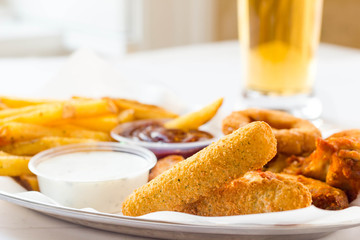 Fastfood with fresh mozzarella sticks, onion rings, french fries and yogurt dip on white table. Served with beer