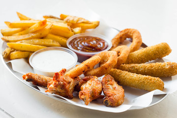 Fastfood plate with mozzarella sticks, chicken wings, onion rings, french fries and dip on white table.