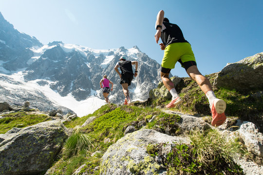 Three trail runners, two men and a woman, running up a steep trail in the mountains in the Alps on a hot, bright summer day.
