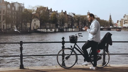 4K Man with bicycle on river bridge with a phone. Casual handsome European bearded male types a message and walks away. - Powered by Adobe