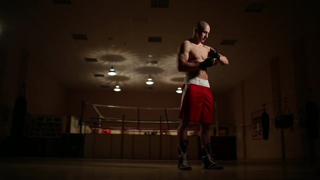A pumped-up man in boxing boots and shorts in the gym against the background of a boxing ring dresses bandages on his hands.