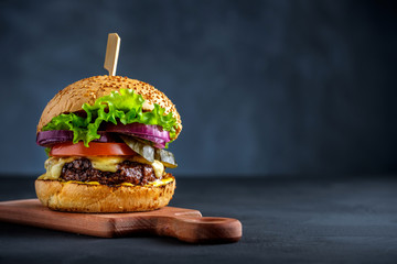 Tasty grilled beef burger with lettuce, cheese and onion served on cutting board on a black wooden table, with copyspace.