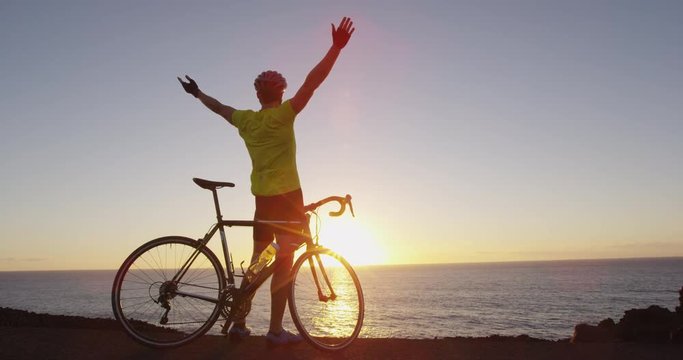 Cycling success and achievement - man cheering reaching goal happy on road bike. Male cyclist on road bike in happy celebration with arms outstretched after biking at sunset. SLOW MOTION RED EPIC.
