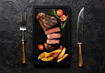Meat Picanha steak, traditional Brazilian cut with tomatoes, potato wedges and rosemary on black meat cutting board. Steak with fork and knife. Black concrete background.