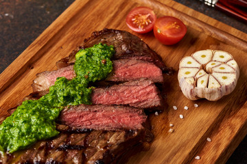 Grilled Black Angus Steak with tomatoes, garlic with chimichurri sauce on meat cutting board.