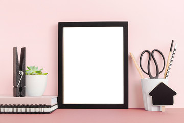 Frame on pink desk, plant and office supplies. Mock up.