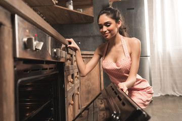 sexy young woman in apron opening oven and smiling at kitchen