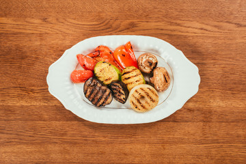 Top view of grilled vegetables and mushrooms on white plate on wooden table