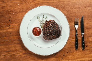 Top view of grilled steak with pepper, rosemary and sauce on white plate on wooden table