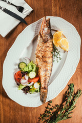 fried fish laying on white plate with vegetables and orange slices over wooden surface with fork and knife on napkin