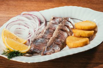 Herring fish pieces with lemon, onion and potato on white plate on wooden table