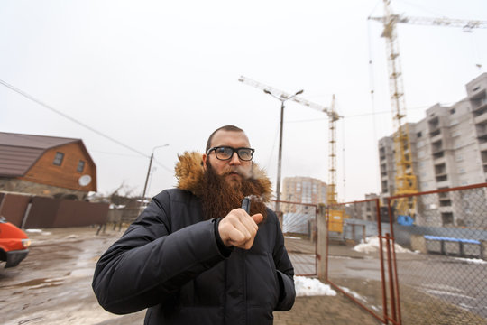 Vape bearded man. Portrait of young guy with large beard in glasses vaping an electronic cigarette opposite the unfinished building of white brick in the winter.