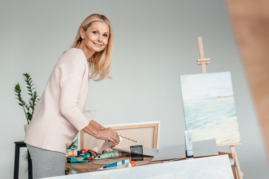 beautiful mature woman smiling at camera while holding art tools in art studio