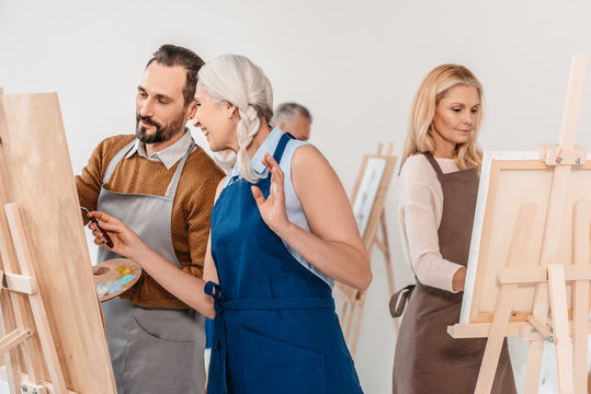 Adult Students In Aprons Painting On Easels During Art Class