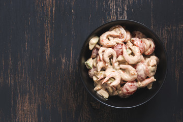 Salad with shrimps, avocado and cherry tomatoes is served in a black bowl on a dark wood background. View from above.