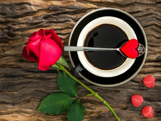 The arrangement for Valentines day of a cup of coffee with a heart shape paper on metal tea spoon and red rose and heart shape candy on wooden slab