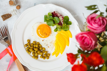 A festive breakfast. Fried egg heart, green peas and salad, a bouquet of roses. View from above. Valentine's Day concept.