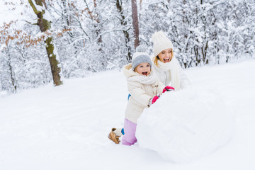 happy mother and cute little daughter making snowman together in winter park