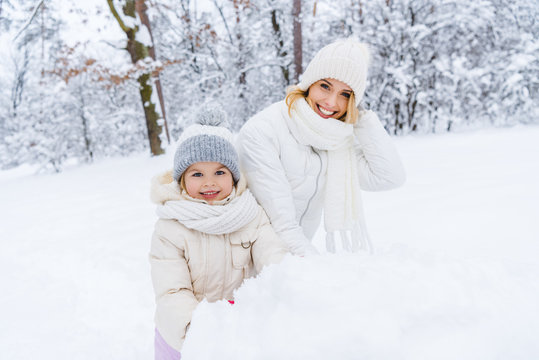 Beautiful Happy Mother And Daughter Smiling At Camera While Making Snowman In Winter Park