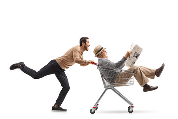 Mature man with a newspaper riding inside a shopping cart being pushed by a young man