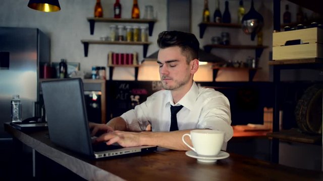 A young man sits in the kitchen at the bar counter in the evening and works in a laptop with important data. Home atmosphere, work in the evening after a hard day with cup of coffee. Home office. 