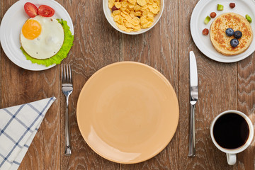 top view of empty brown plate,  pancake with blueberries and fried eggs with tomatoes and lettuce on a plates, cornflakes