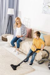 happy pregnant woman sitting on sofa and looking at cute little boy cleaning carpet with vacuum cleaner