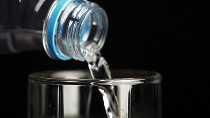 Water poured from the bottle into a transparent glass cup on a black background. Water is poured into a glass from bottle, close up