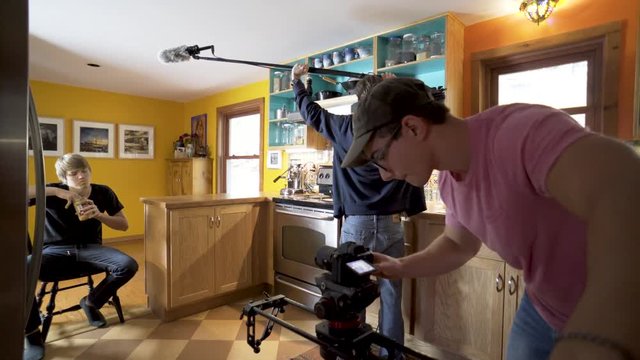 Teen filmmaker slides the camera forward while audio engineer holds the sound boom and teen actor acts his part.