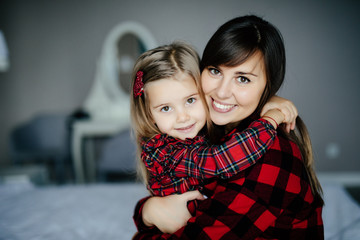 Beautiful young mom and her little daughter are cuddling, smiling while sitting on bed at home