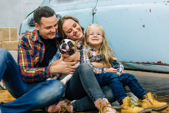 Young happy family in checked shirts sittng near blue retro car with dog. Couple in love with cute daughter having fun with french bulldog