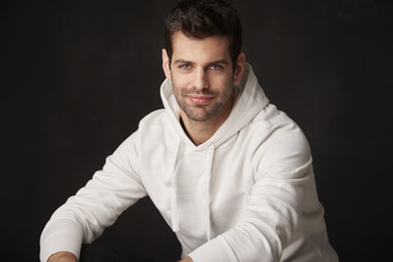 Studio shot of a handsome young man sitting at dark background while looking at camera and smiling.