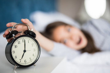 Portrait of shocked cute little girl holding alarm clock and looking at camera while lying in bed in the morning