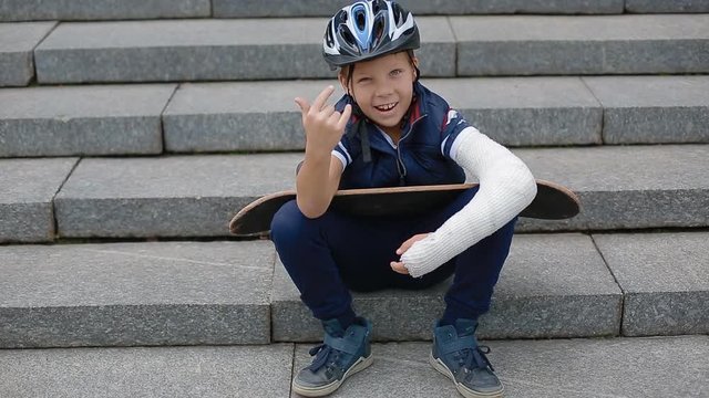 White boy with broken arm makng the rock sign sitting on the granite stairs with his skateboard.