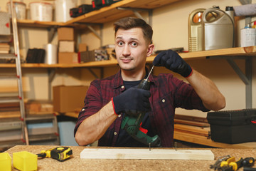 Handsome smiling caucasian young man in plaid shirt, black T-shirt, gloves drilling with power drill in piece of wood, working in carpentry workshop at wooden table place with different tools.