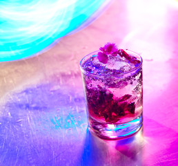 Vibrant cranberry cocktail garnished with edible blossom, set against a textured metallic background with neon highlights — perfect for nightlife and club vibes.