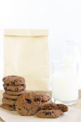 Chocolate chip cookies on brown paper and sack cloth with glass of milk and paper bag on white background.