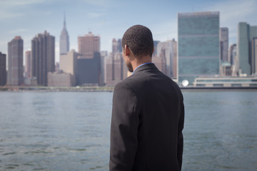 Fototapeta na wymiar Back view of African American male professional in suits standing across New York City skyline and contemplating