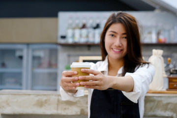 Young asia woman barista holding a diaposable coffee cup with smiling face at cafe counter background, small business owner, food and drink industry concept