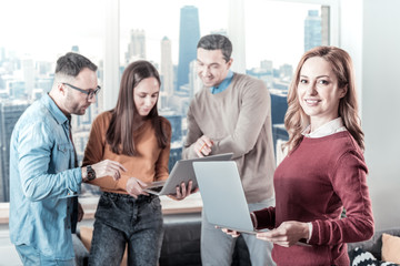 Best work. Confident pretty beautiful woman standing near her colleagues holding the laptop and looking straight.