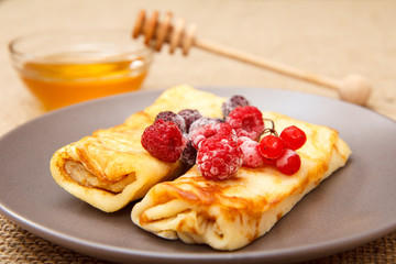 Homemade pancakes filled with cottage cheese and topped with frozen raspberries and blackberries on plate and honey in glass bowl with wooden spoon