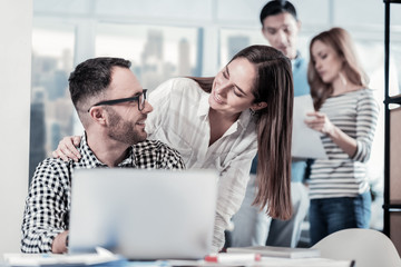 Well done. Bespectacled joyful unshaken man sitting in the office opposite laptop looking at the colleague and smiling.