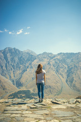 Fototapeta premium A young girl stands on the edge of a cliff. Mountain in the background. Soldiers stand. Blue sky. Young in jeans and a t-shirt