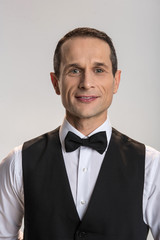 Adorable tie bow. Portrait of  handsome energetic male waiter posing on the grey background and  gazing  at the camera while smiling