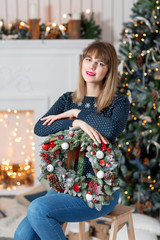 Young woman holding a Christmas wreath with fir branches for the holiday. The new year celebration. on the background of fireplace and Christmas tree, lights, garlands