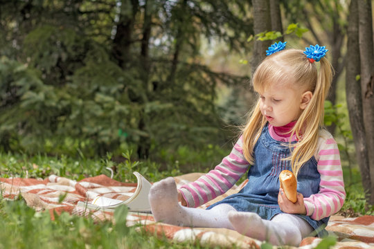 Cute little blonde girl with two ponytails relaxing with a book and a bun in the city park on a spring sunny day.