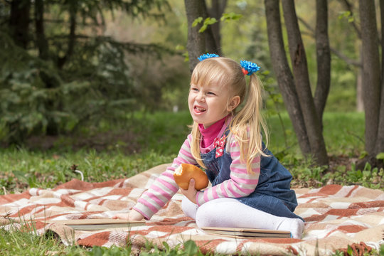 Cute little blonde girl with two ponytails relaxing with a book and a bun in the city park on a spring sunny day.