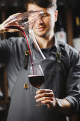 Bartender pours red wine in glass from big transparent vessel