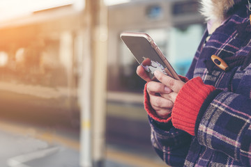 Dried hand of middle age woman in winter coat with a cell phone in her hand waiting on the platform of a railway station for train to arrive. Public transport.