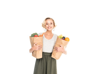 young woman holding paper bags with fruits and vegetables and smiling at camera isolated on white